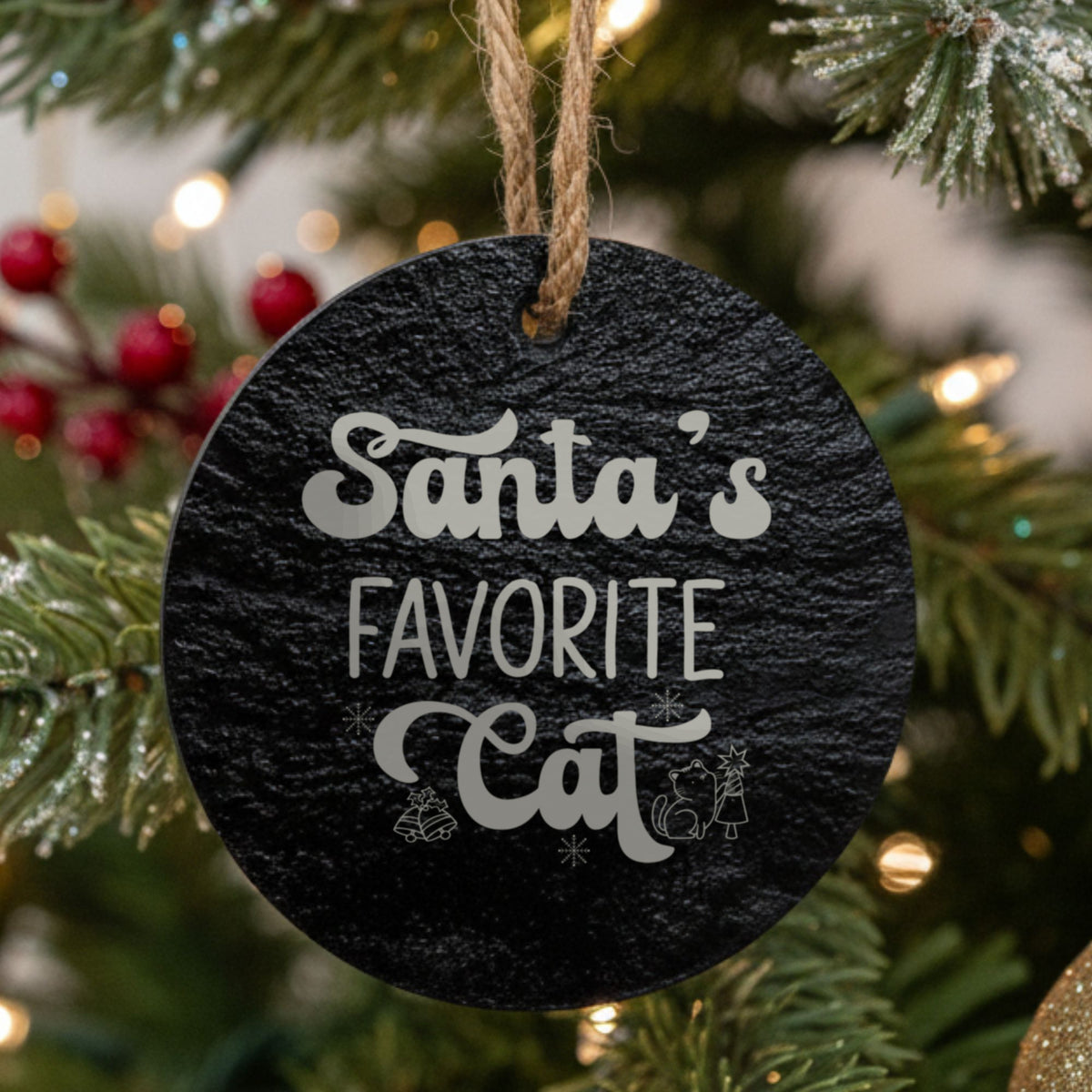 Round black slate Christmas ornament hanging from a jute string on a decorated Christmas tree, laser engraved in white with “Santas Favorite Cat”.