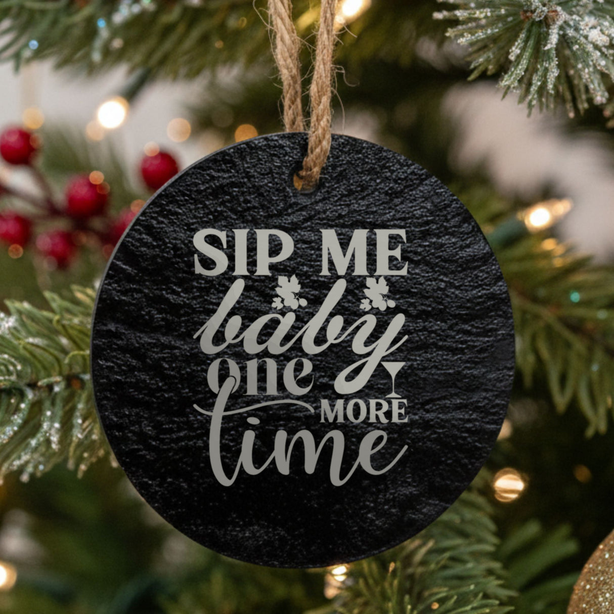 Round black slate Christmas ornament hanging from a jute string on a decorated Christmas tree, laser engraved in white with “Sip Me Baby One More Time”.