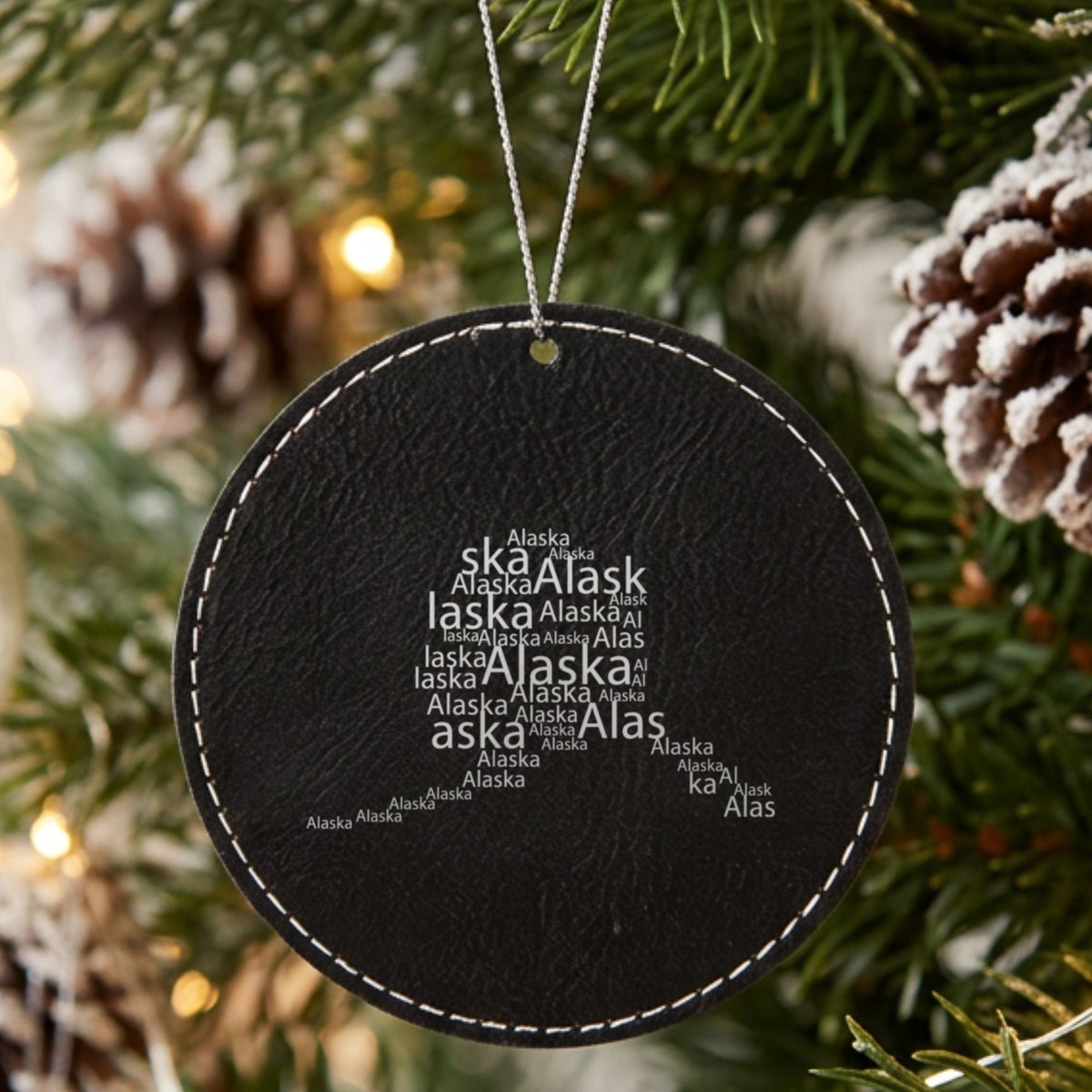 Black leatherette round Christmas ornament laser engraved with the Alaska Word Cloud design, hanging on a tree