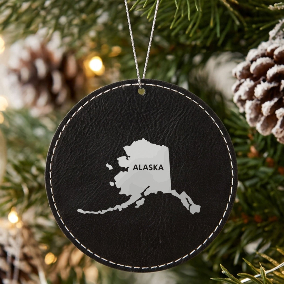 Black leatherette round Christmas ornament laser engraved with the Alaska Bold State Name design, hanging on a tree