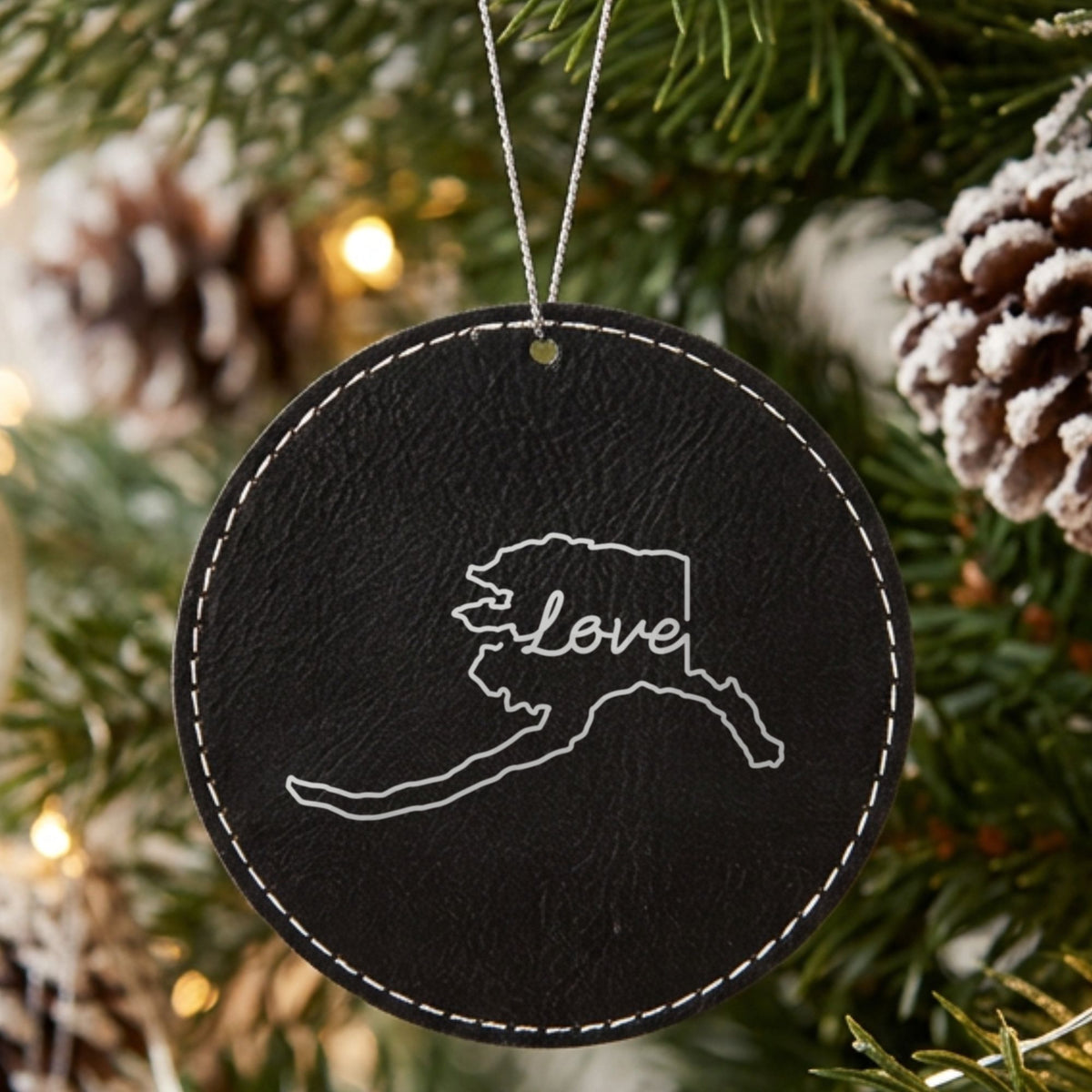 Black leatherette round Christmas ornament laser engraved with the Alaska Hand-Drawn Love design, hanging on a tree