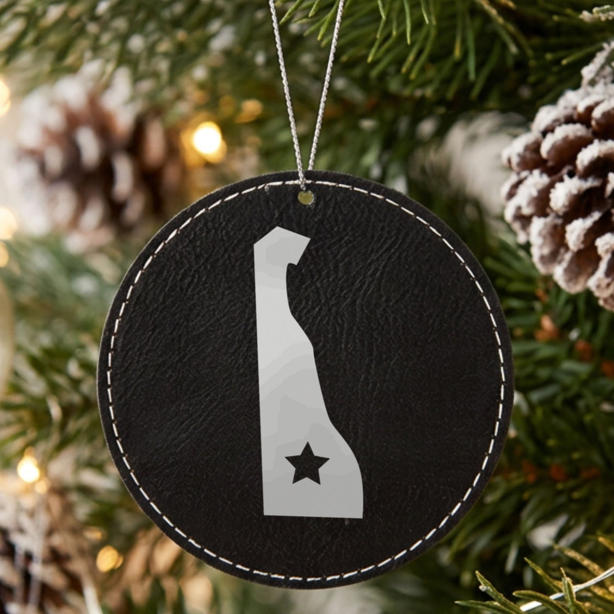 Black leatherette round Christmas ornament laser engraved with the Delaware Star Accent design, hanging on a tree