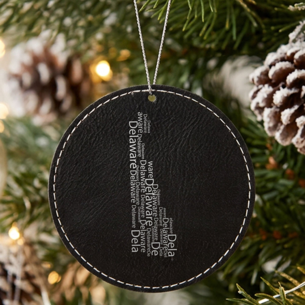 Black leatherette round Christmas ornament laser engraved with the Delaware Word Cloud design, hanging on a tree
