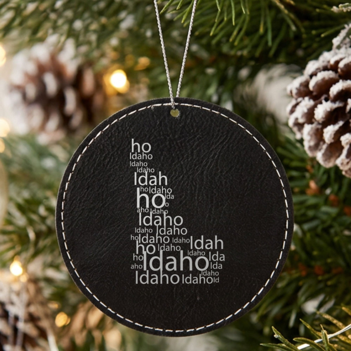 Black leatherette round Christmas ornament laser engraved with the Idaho Word Cloud design, hanging on a tree