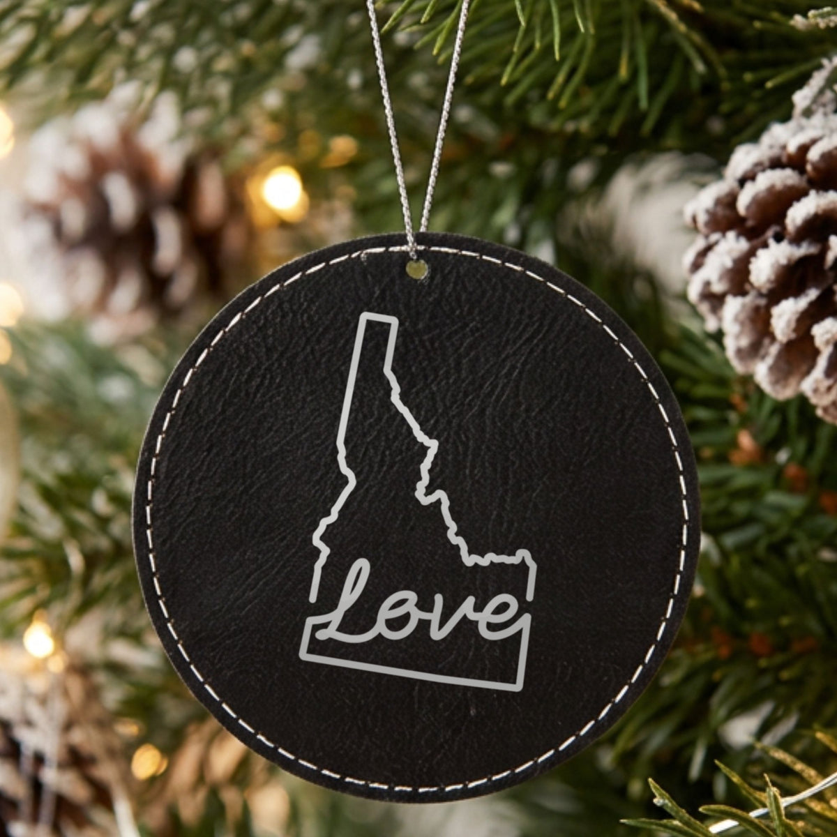 Black leatherette round Christmas ornament laser engraved with the Idaho Hand-Drawn Love design, hanging on a tree
