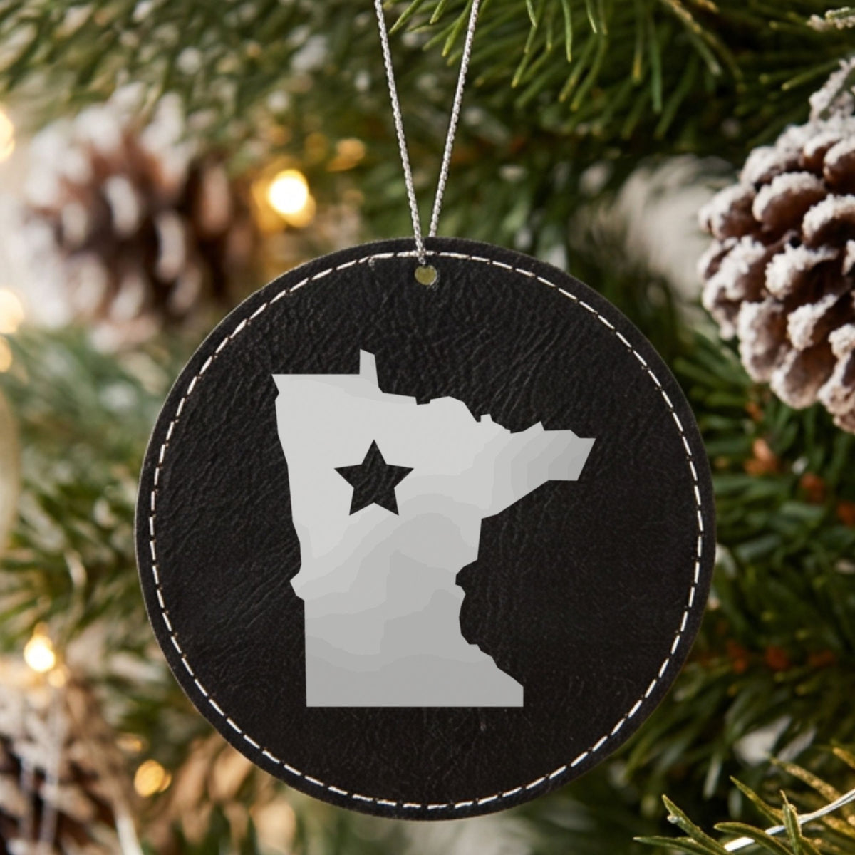 Black leatherette round Christmas ornament laser engraved with the Minnesota Star Accent design, hanging on a tree