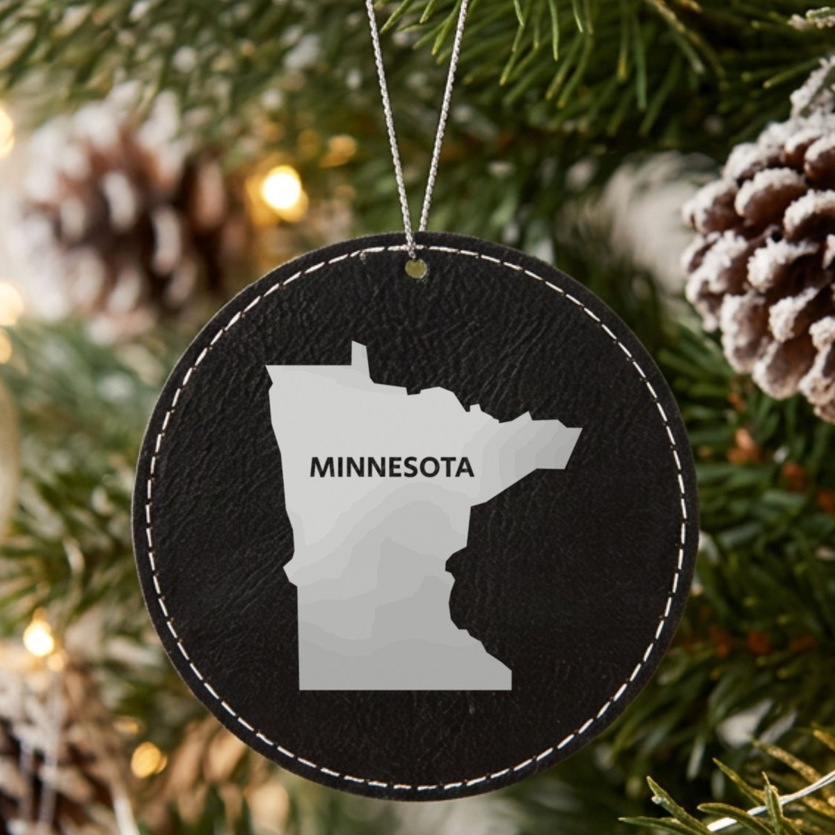 Black leatherette round Christmas ornament laser engraved with the Minnesota Bold State Name design, hanging on a tree
