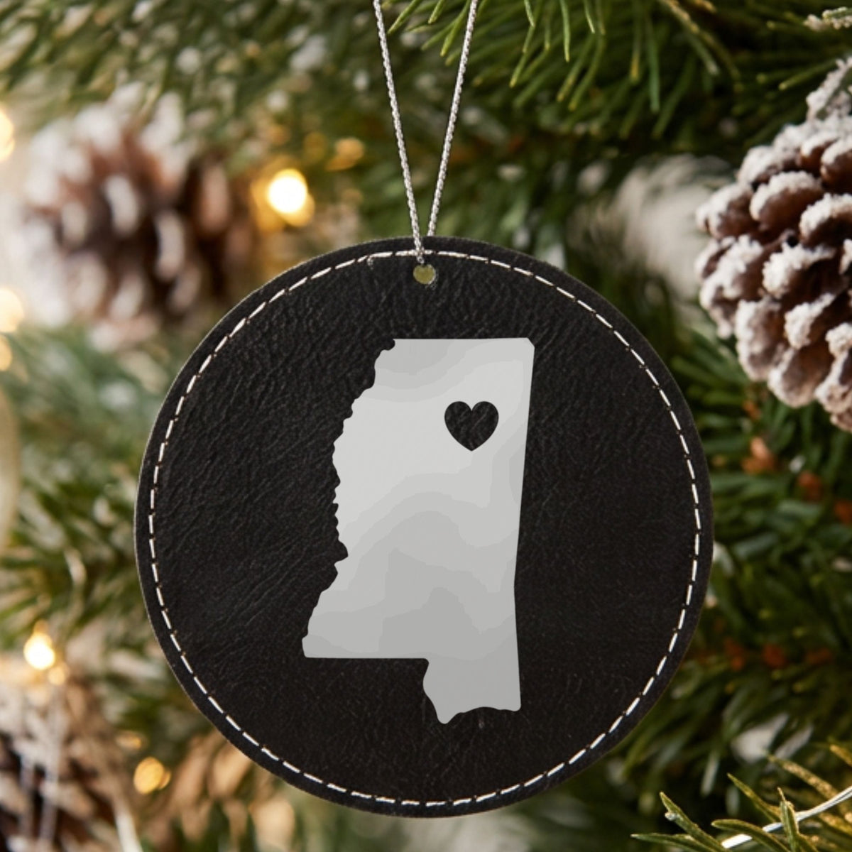 Black leatherette round Christmas ornament laser engraved with the Mississippi Heart Accent design, hanging on a tree