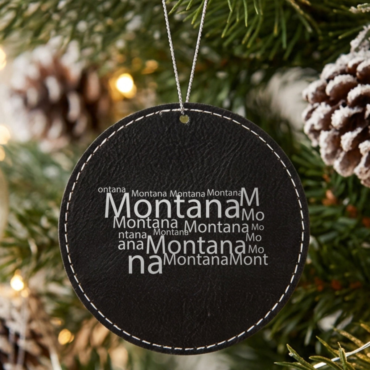 Black leatherette round Christmas ornament laser engraved with the Montana Word Cloud design, hanging on a tree