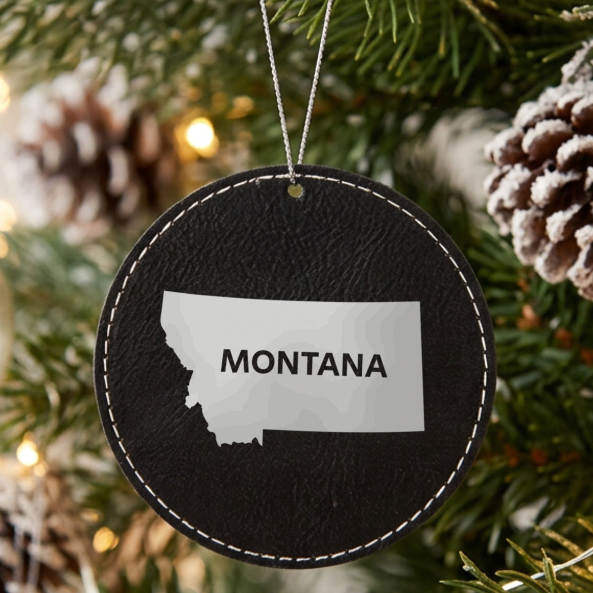 Black leatherette round Christmas ornament laser engraved with the Montana Bold State Name design, hanging on a tree