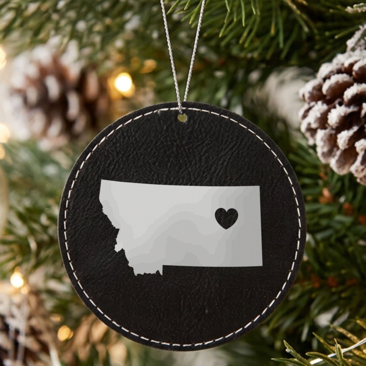 Black leatherette round Christmas ornament laser engraved with the Montana Heart Accent design, hanging on a tree