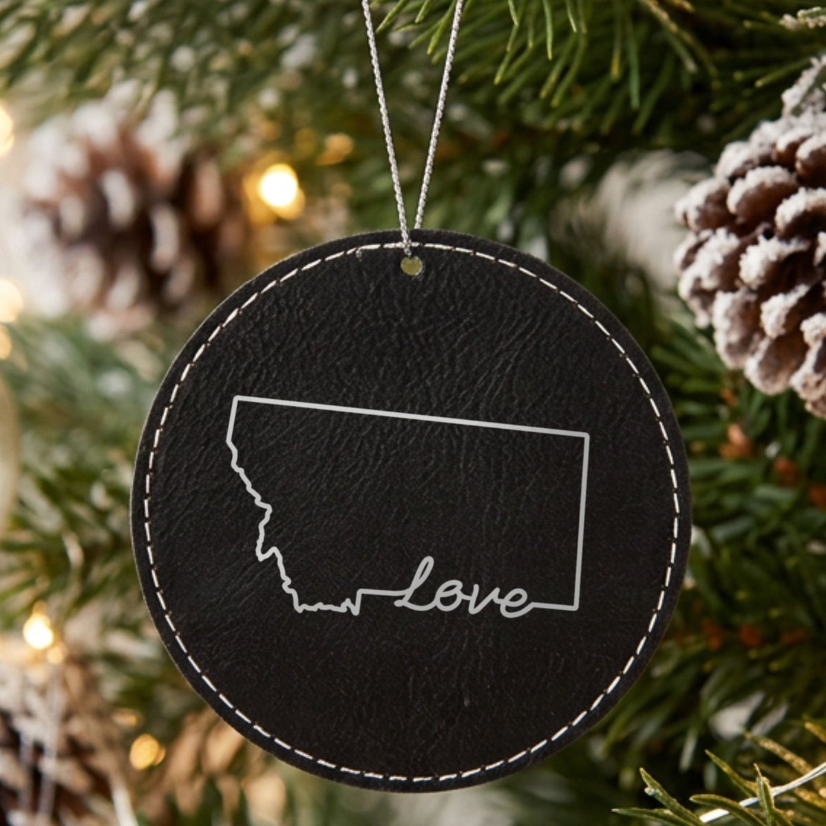 Black leatherette round Christmas ornament laser engraved with the Montana Hand-Drawn Love design, hanging on a tree