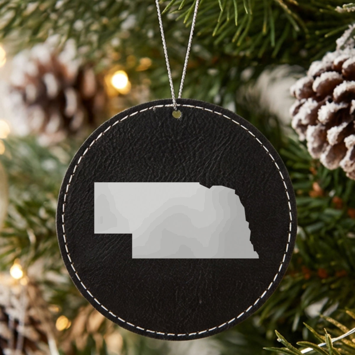 Black leatherette round Christmas ornament laser engraved with the Nebraska Engraved State design, hanging on a tree