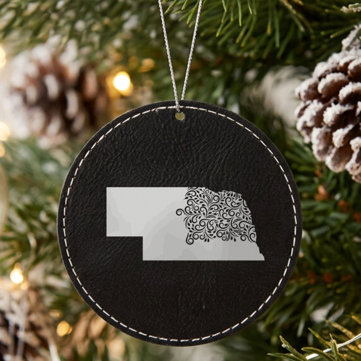 Black leatherette round Christmas ornament laser engraved with the Nebraska Floral Damask design, hanging on a tree