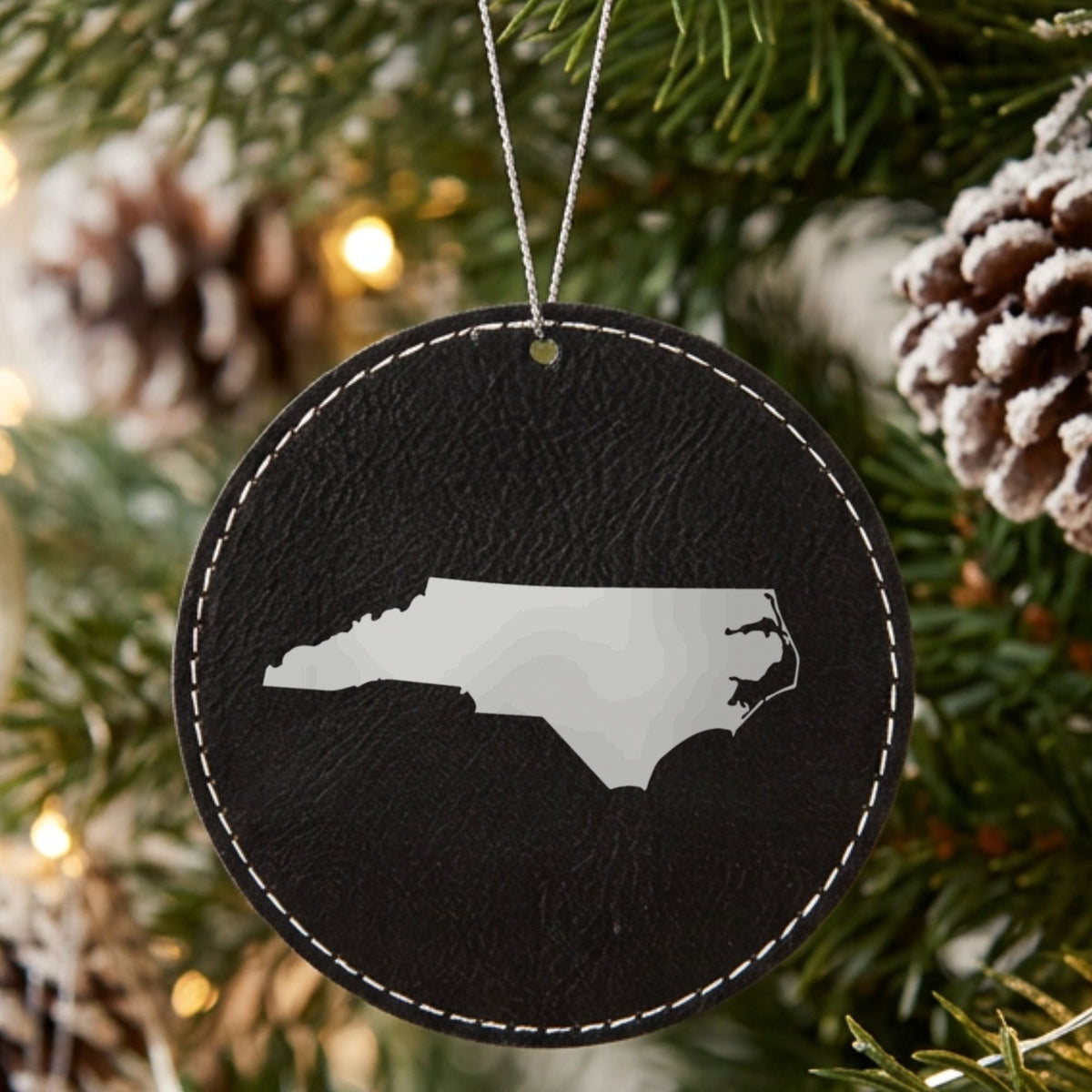 Black leatherette round Christmas ornament laser engraved with the North Carolina Engraved State design, hanging on a tree
