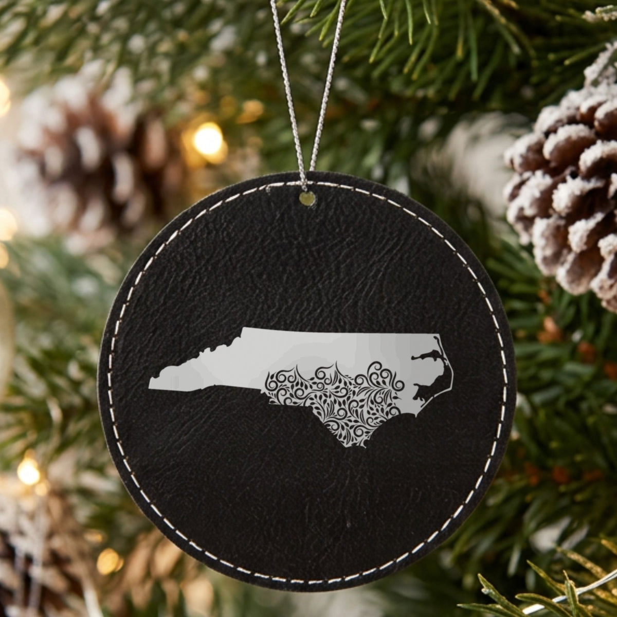 Black leatherette round Christmas ornament laser engraved with the North Carolina Floral Damask design, hanging on a tree