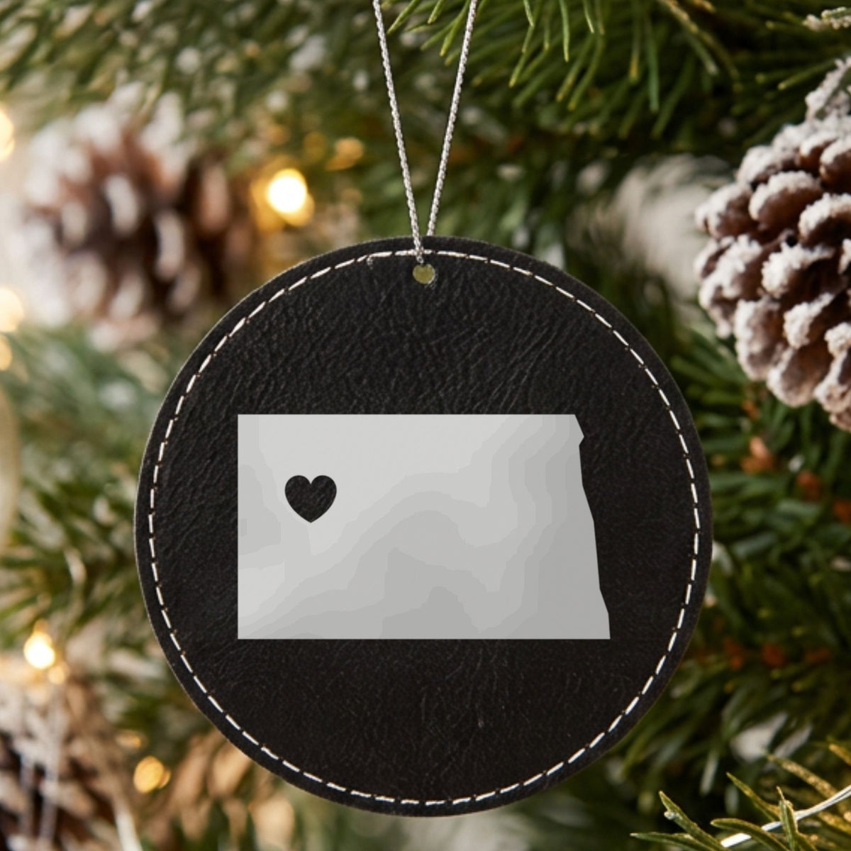 Black leatherette round Christmas ornament laser engraved with the North Dakota Heart Accent design, hanging on a tree