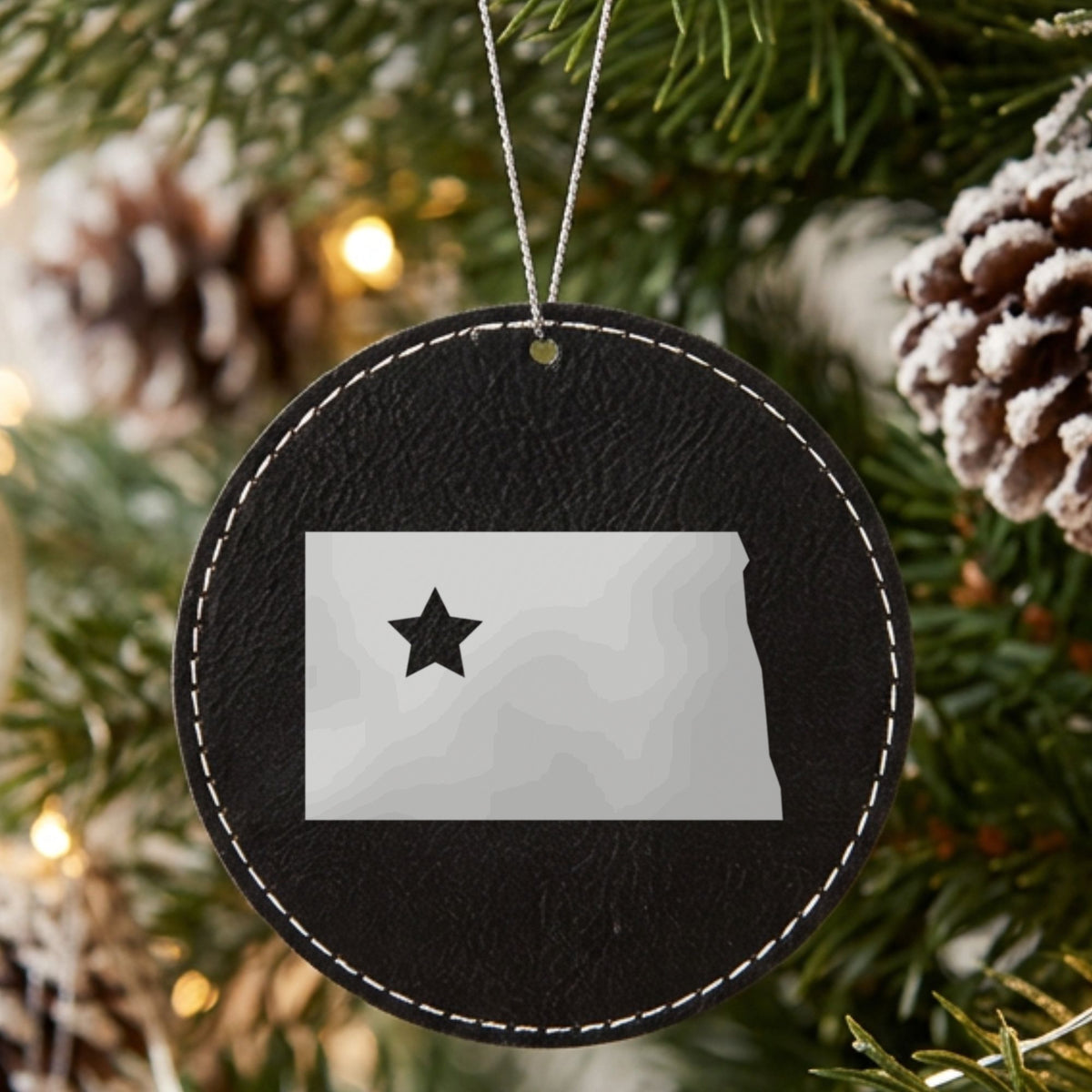 Black leatherette round Christmas ornament laser engraved with the North Dakota Star Accent design, hanging on a tree