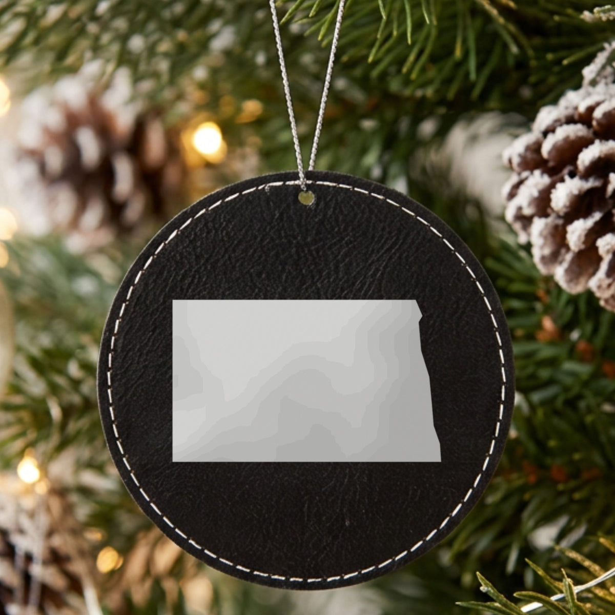 Black leatherette round Christmas ornament laser engraved with the North Dakota Engraved State design, hanging on a tree