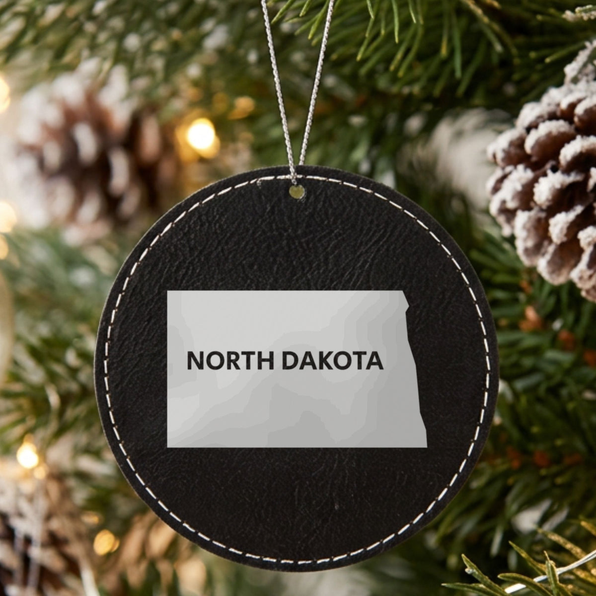 Black leatherette round Christmas ornament laser engraved with the North Dakota Bold State Name design, hanging on a tree