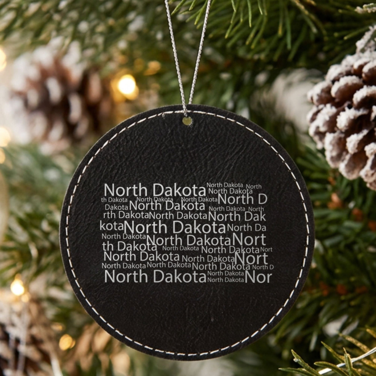 Black leatherette round Christmas ornament laser engraved with the North Dakota Word Cloud design, hanging on a tree