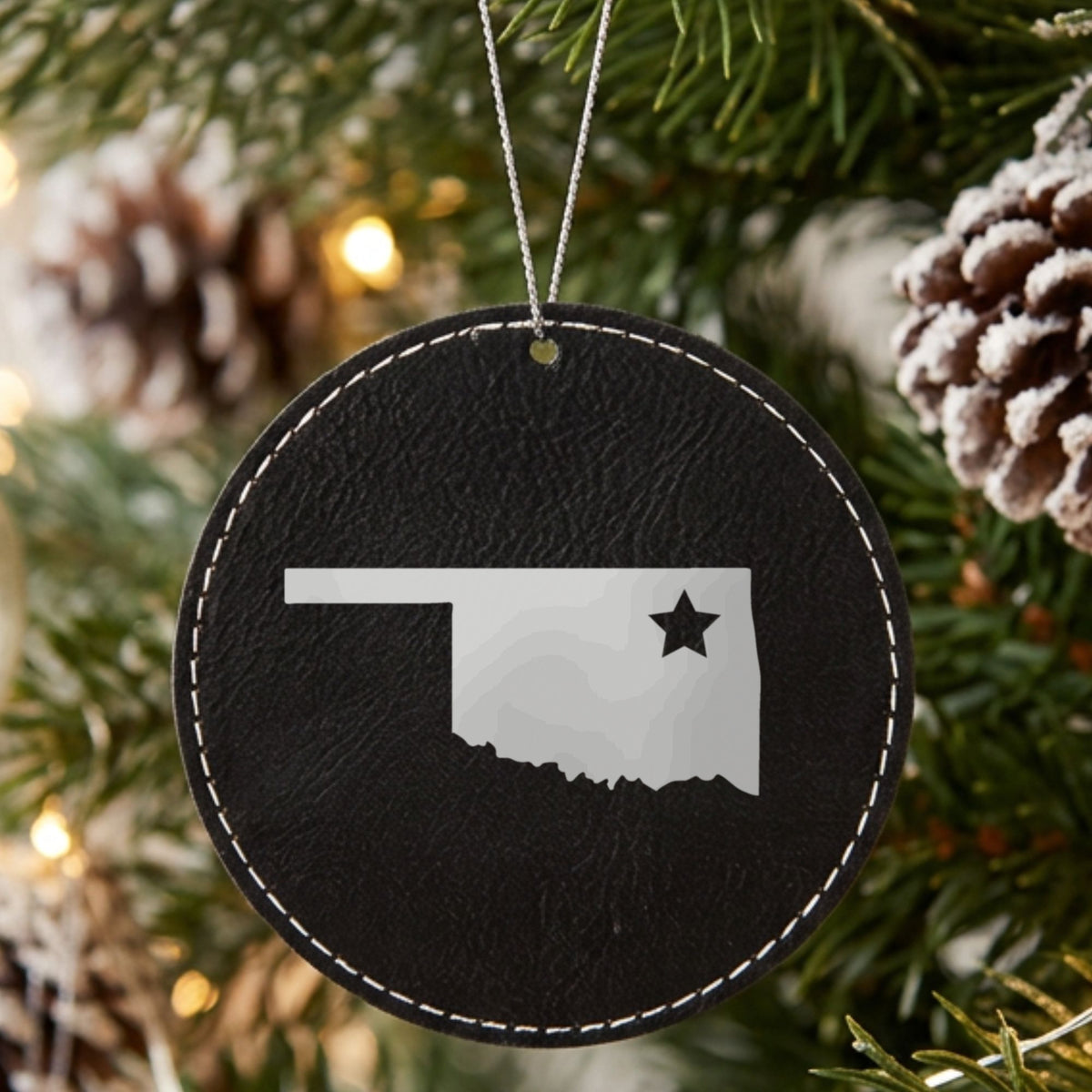 Black leatherette round Christmas ornament laser engraved with the Oklahoma Star Accent design, hanging on a tree