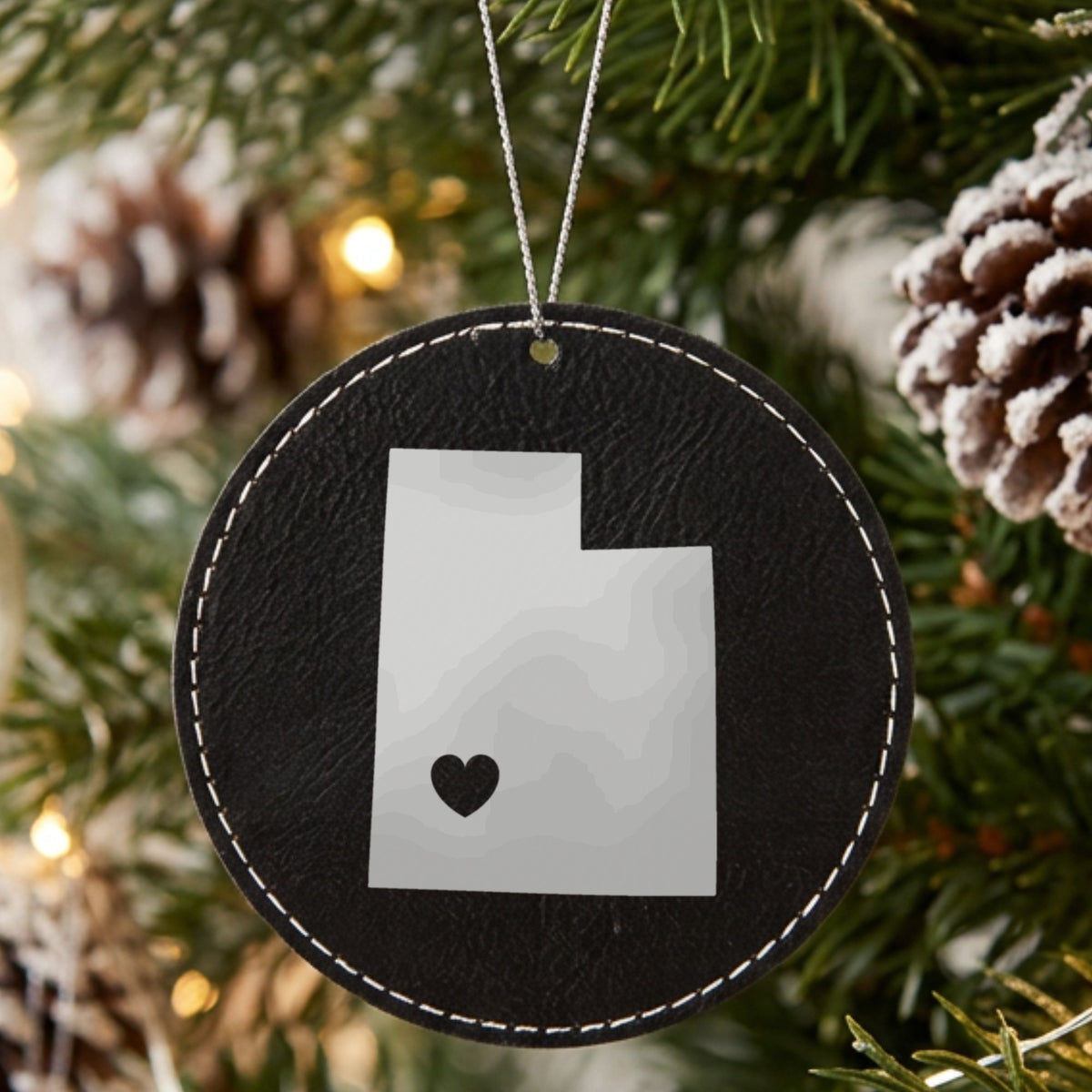 Black leatherette round Christmas ornament laser engraved with the Utah Heart Accent design, hanging on a tree