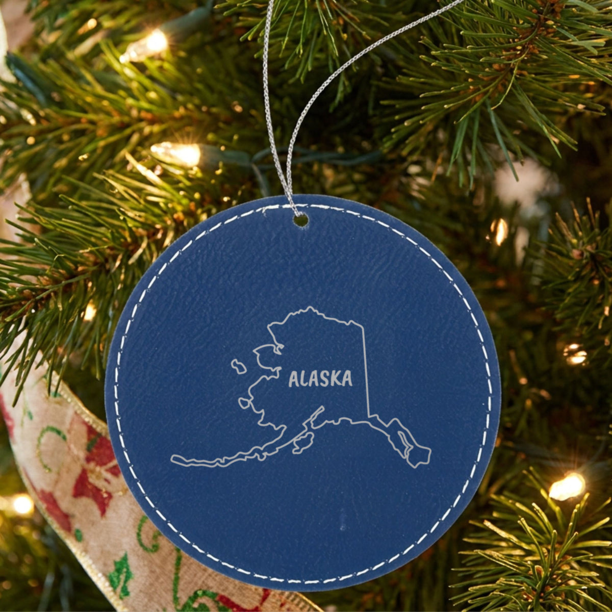 Blue leatherette round Christmas ornament laser engraved with the Alaska Brush Lettering design, hanging on a tree