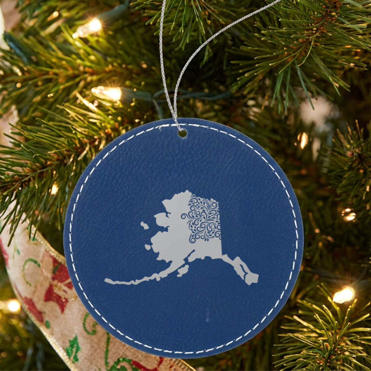 Blue leatherette round Christmas ornament laser engraved with the Alaska Floral Damask design, hanging on a tree
