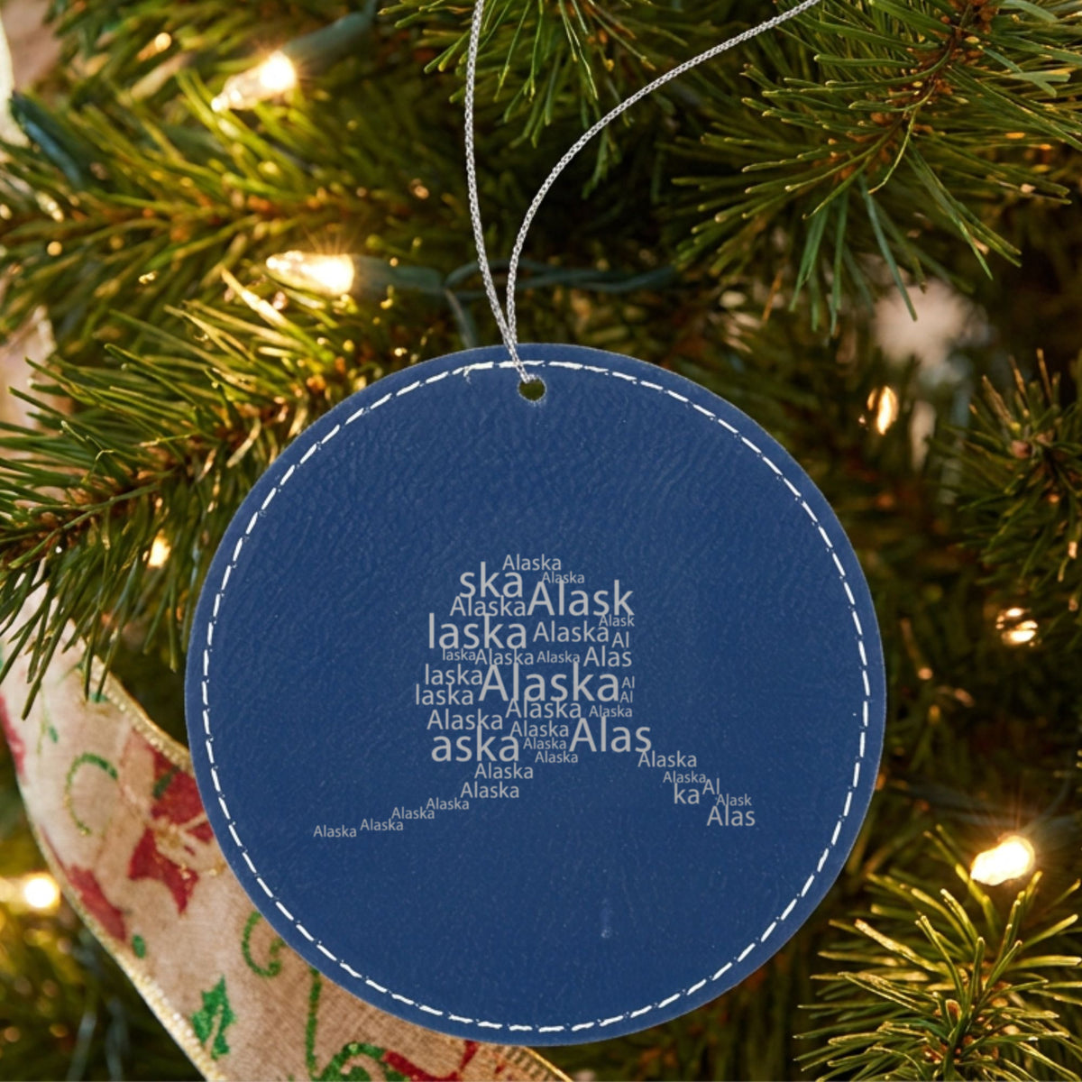 Blue leatherette round Christmas ornament laser engraved with the Alaska Word Cloud design, hanging on a tree
