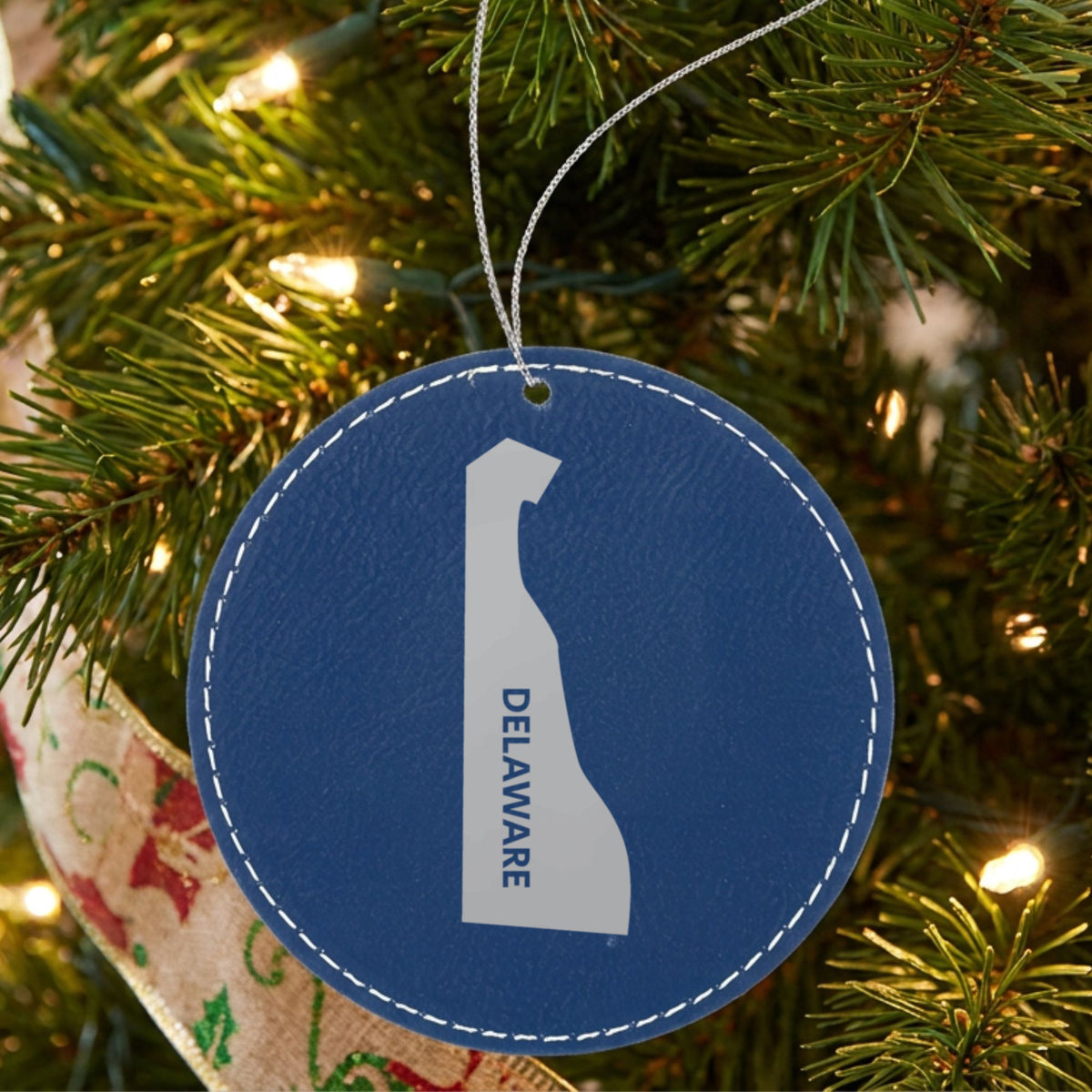 Blue leatherette round Christmas ornament laser engraved with the Delaware Vertical Name (Engraved) design, hanging on a tree