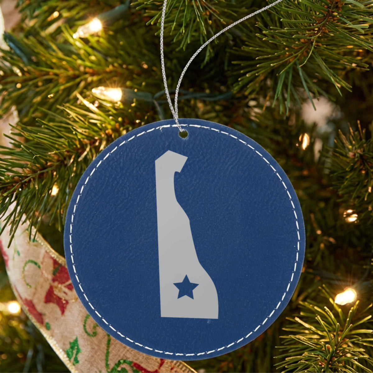 Blue leatherette round Christmas ornament laser engraved with the Delaware Star Accent design, hanging on a tree