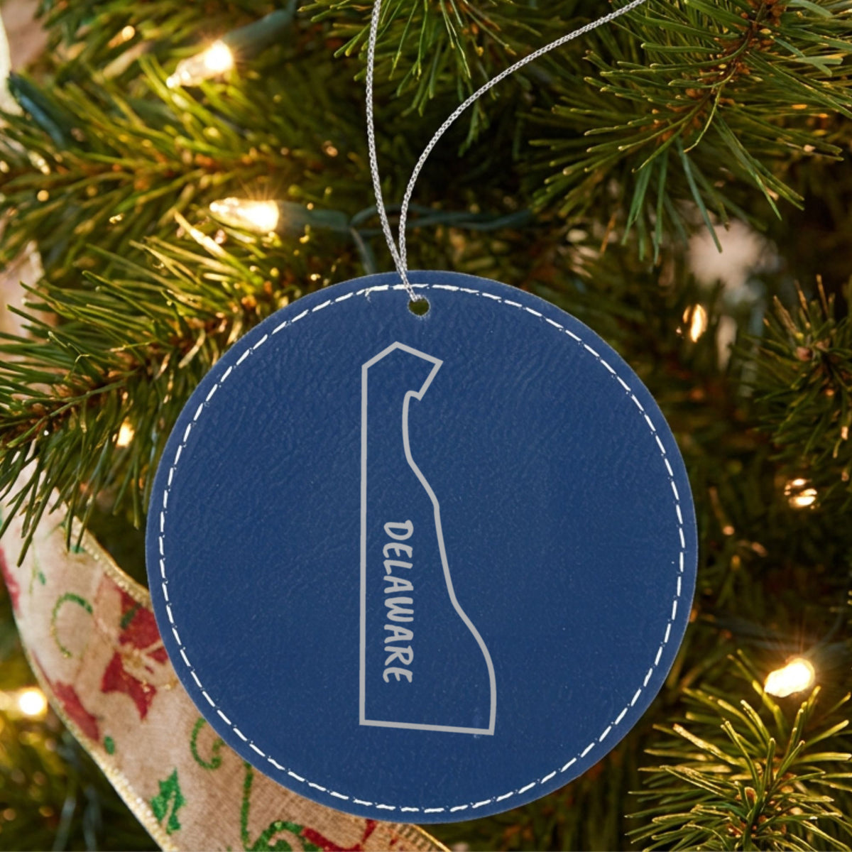 Blue leatherette round Christmas ornament laser engraved with the Delaware Vertical Name (Outline) design, hanging on a tree