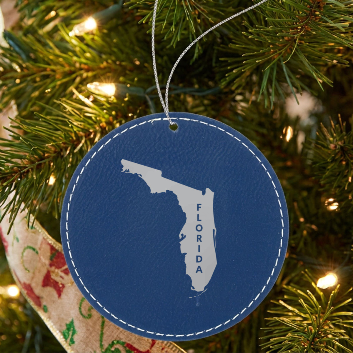 Blue leatherette round Christmas ornament laser engraved with the Florida Vertical Name (Engraved) design, hanging on a tree