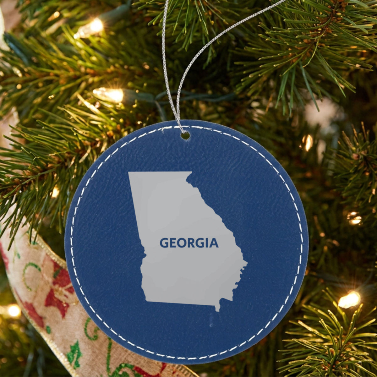 Blue leatherette round Christmas ornament laser engraved with the Georgia Bold State Name design, hanging on a tree