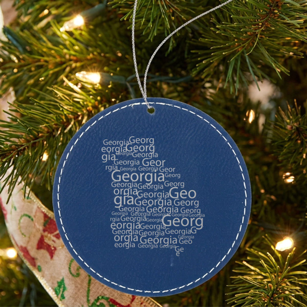 Blue leatherette round Christmas ornament laser engraved with the Georgia Word Cloud design, hanging on a tree