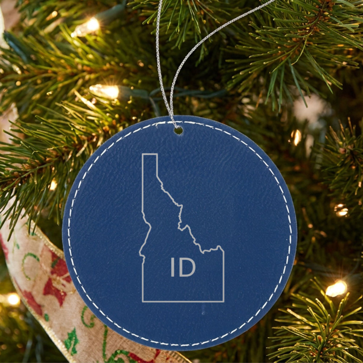 Blue leatherette round Christmas ornament laser engraved with the Idaho ID Initials design, hanging on a tree