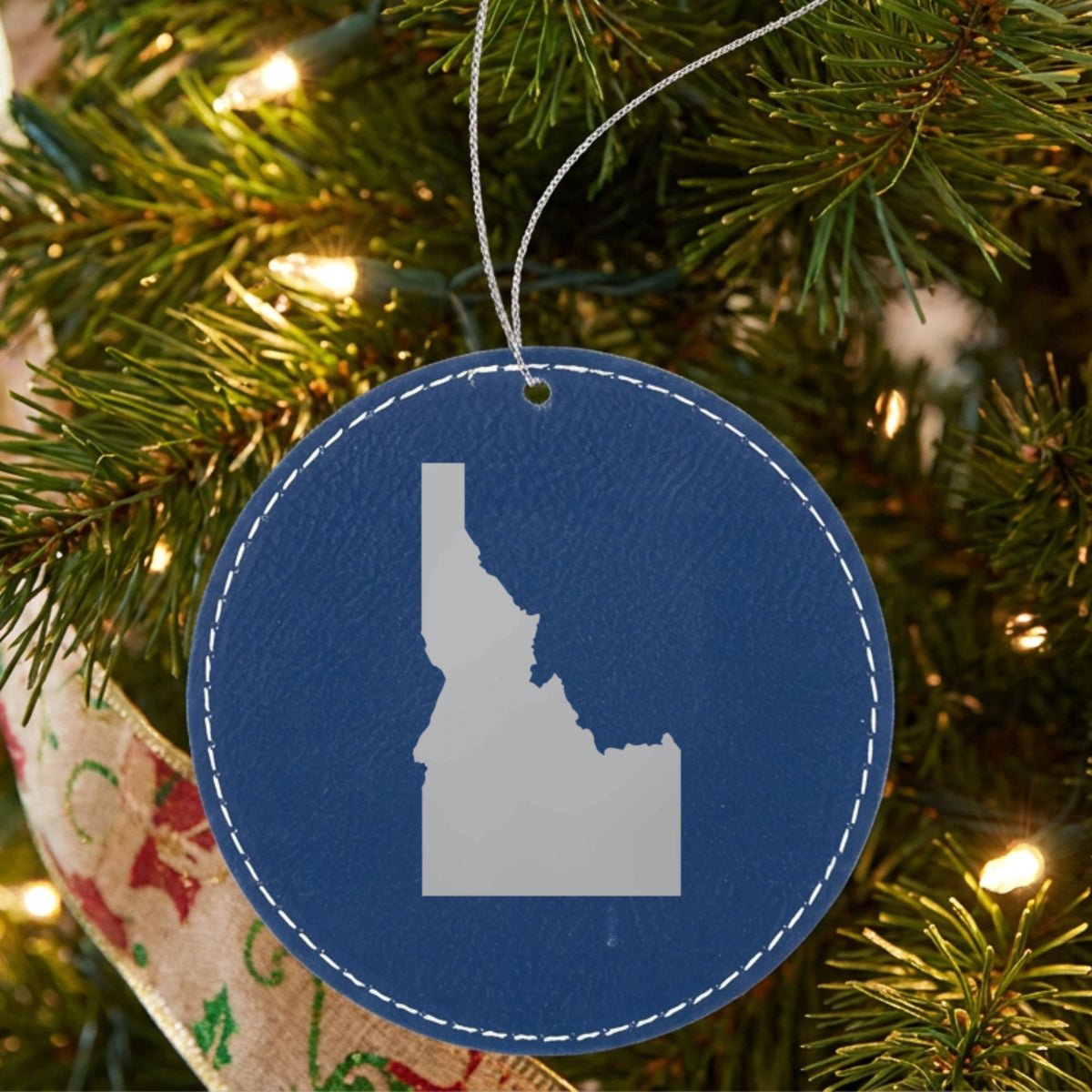 Blue leatherette round Christmas ornament laser engraved with the Idaho Engraved State design, hanging on a tree