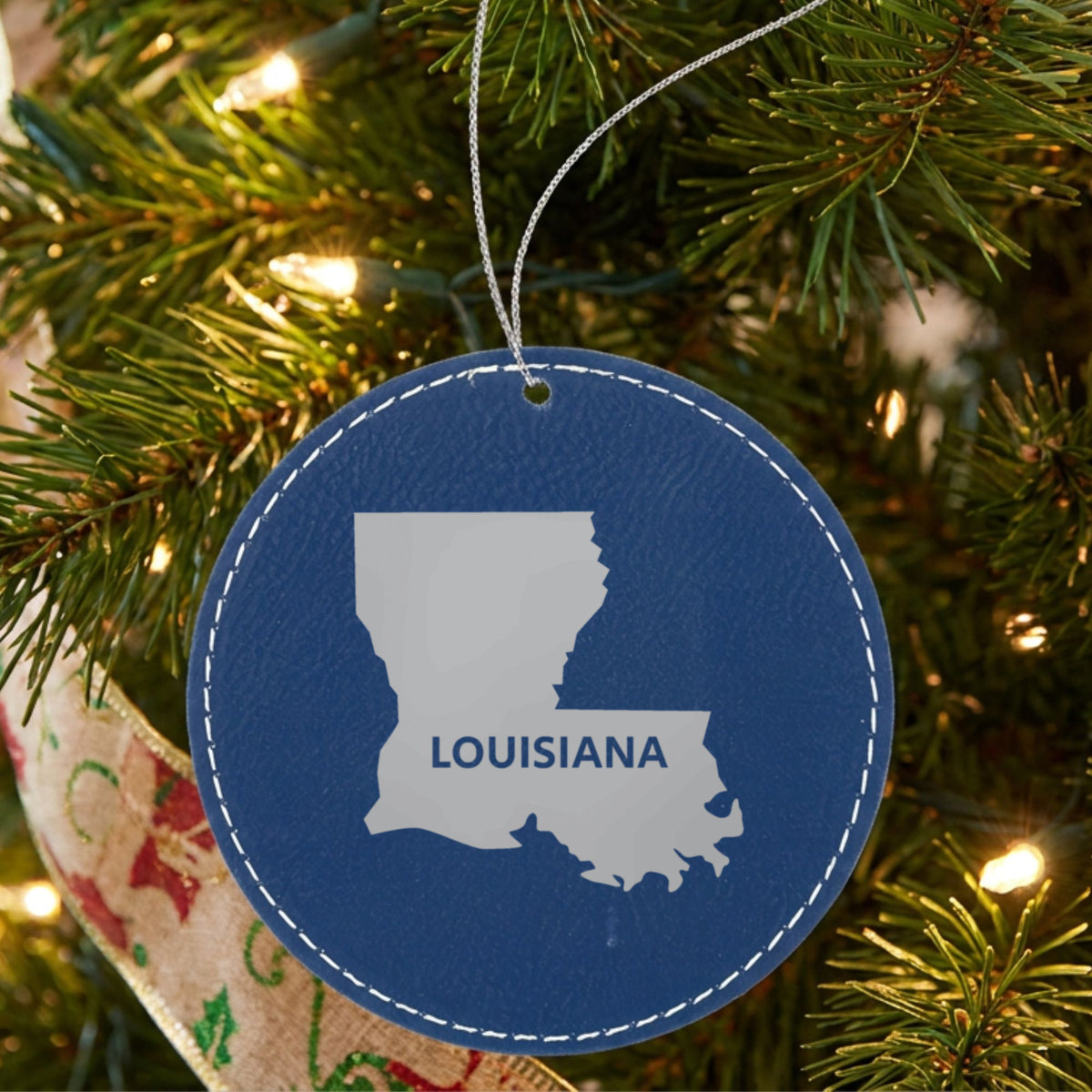 Blue leatherette round Christmas ornament laser engraved with the Louisiana Bold State Name design, hanging on a tree