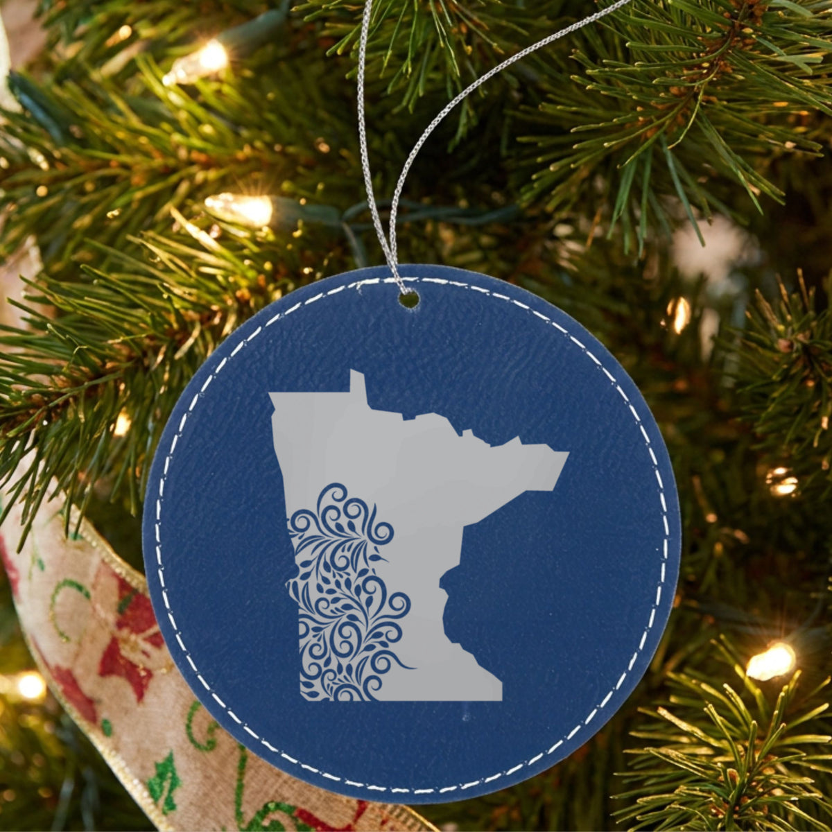 Blue leatherette round Christmas ornament laser engraved with the Minnesota Floral Damask design, hanging on a tree