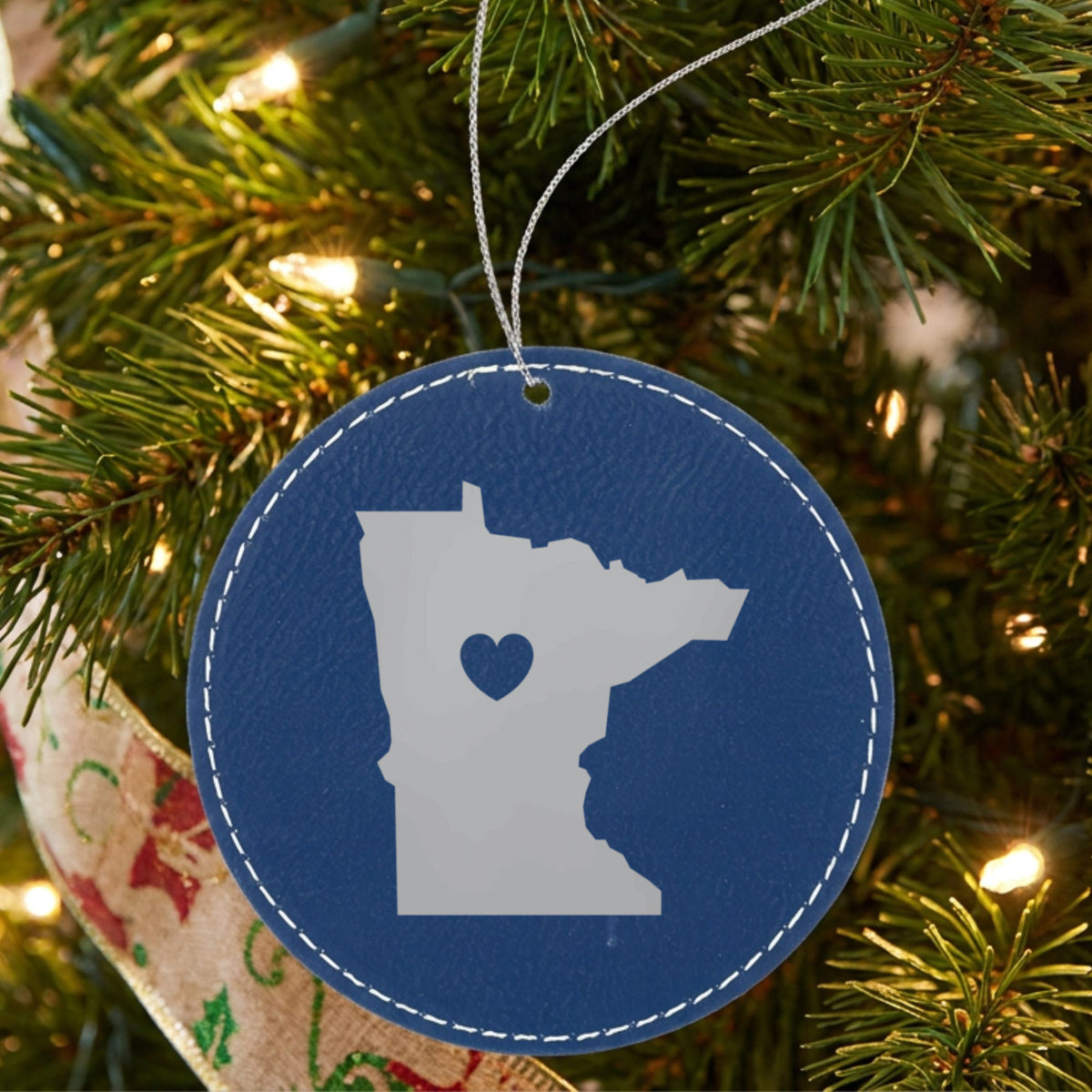 Blue leatherette round Christmas ornament laser engraved with the Minnesota Heart Accent design, hanging on a tree