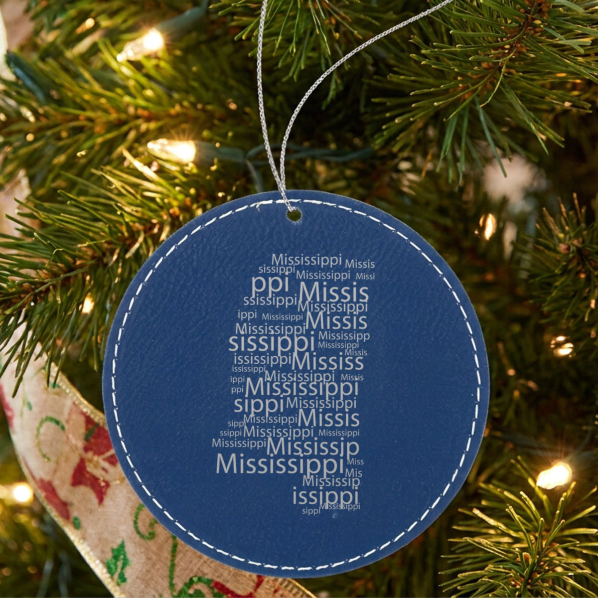 Blue leatherette round Christmas ornament laser engraved with the Mississippi Word Cloud design, hanging on a tree