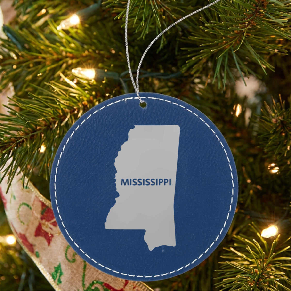 Blue leatherette round Christmas ornament laser engraved with the Mississippi Bold State Name design, hanging on a tree