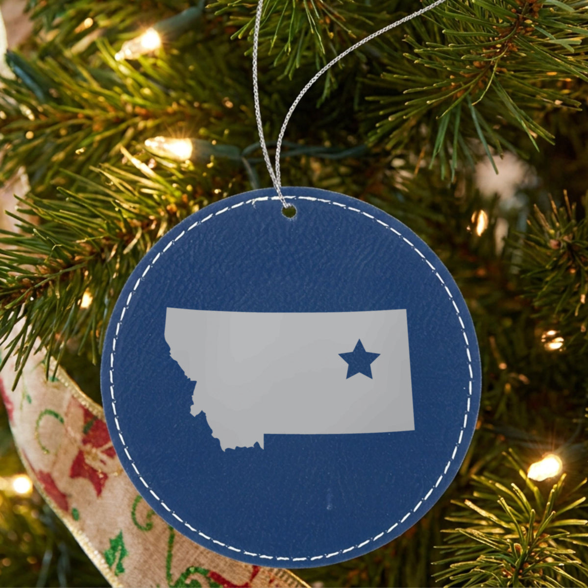 Blue leatherette round Christmas ornament laser engraved with the Montana Star Accent design, hanging on a tree