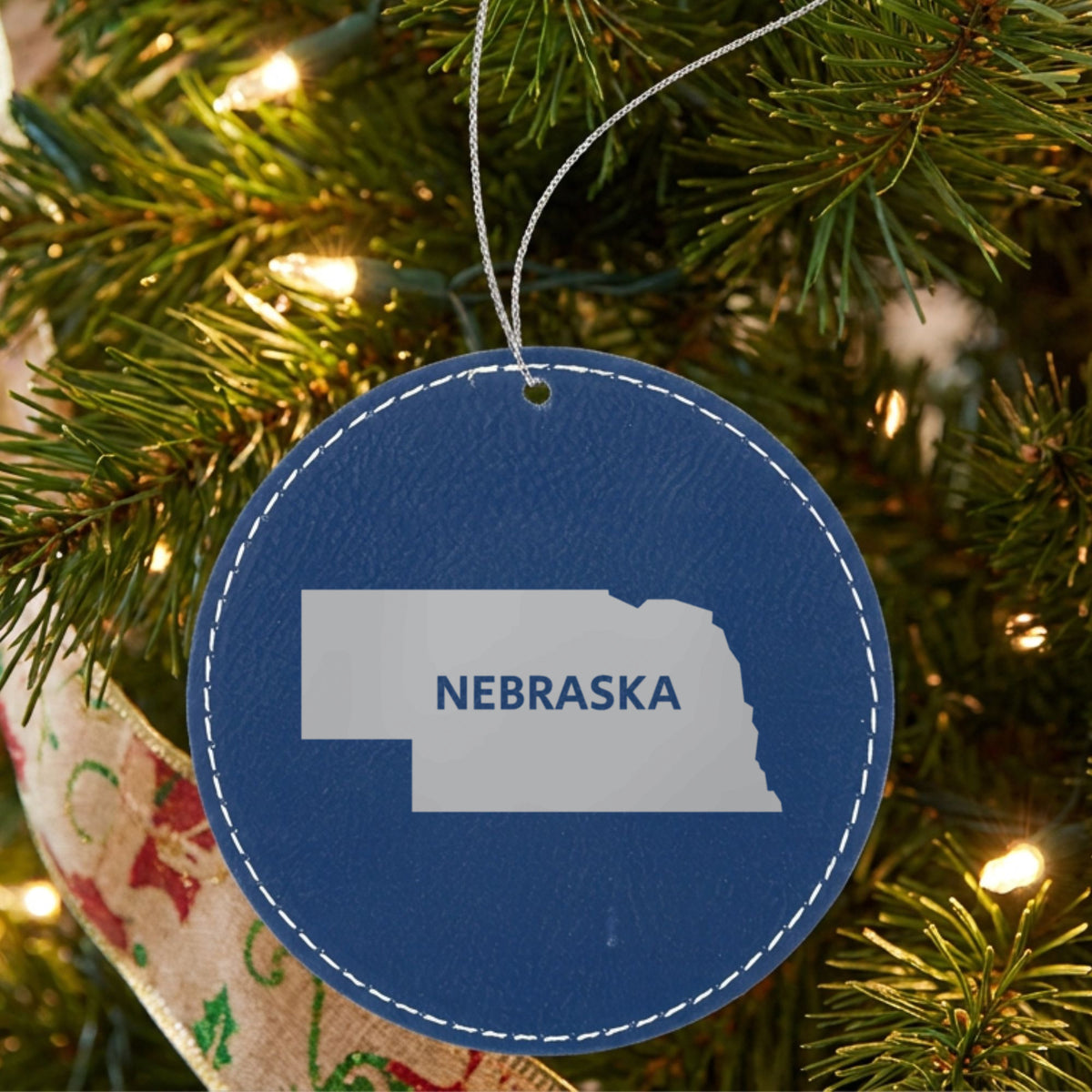 Blue leatherette round Christmas ornament laser engraved with the Nebraska Bold State Name design, hanging on a tree