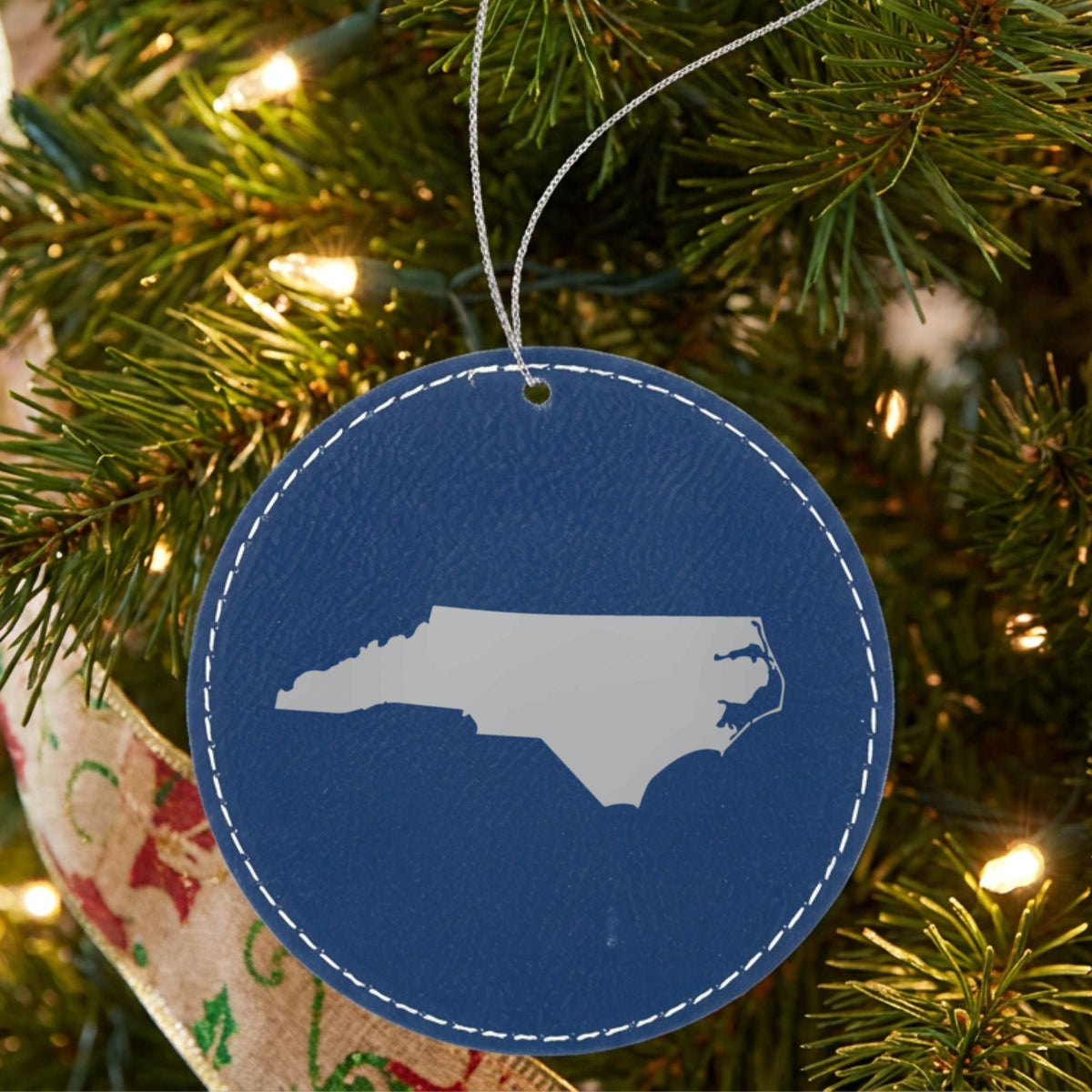 Blue leatherette round Christmas ornament laser engraved with the North Carolina Engraved State design, hanging on a tree