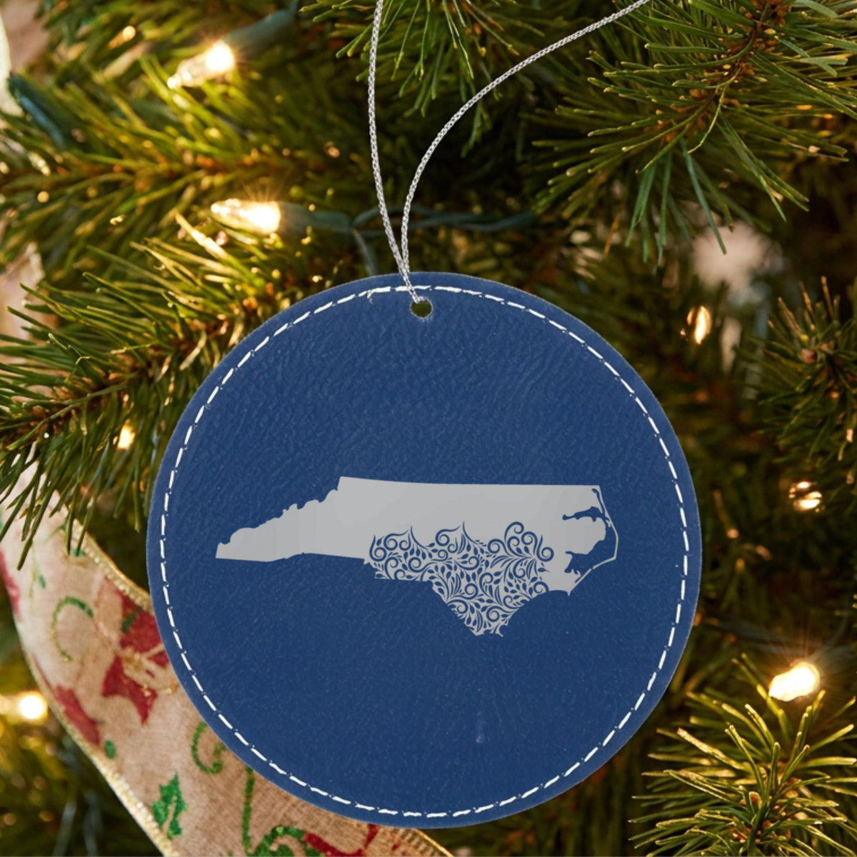 Blue leatherette round Christmas ornament laser engraved with the North Carolina Floral Damask design, hanging on a tree