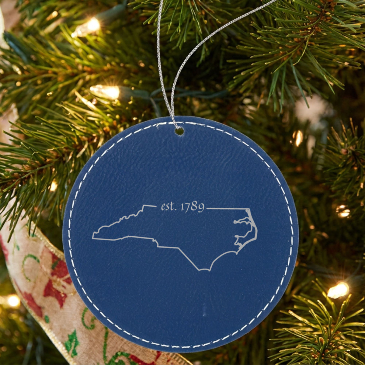 Blue leatherette round Christmas ornament laser engraved with the North Carolina Est. 1789 design, hanging on a tree