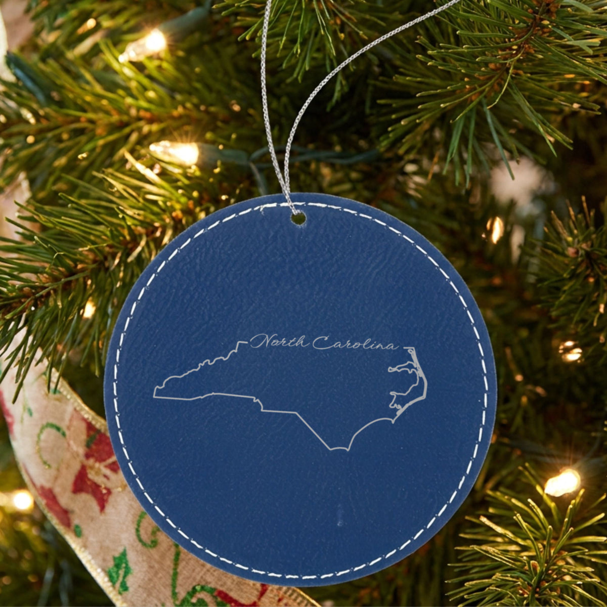 Blue leatherette round Christmas ornament laser engraved with the North Carolina North Carolina Script design, hanging on a tree