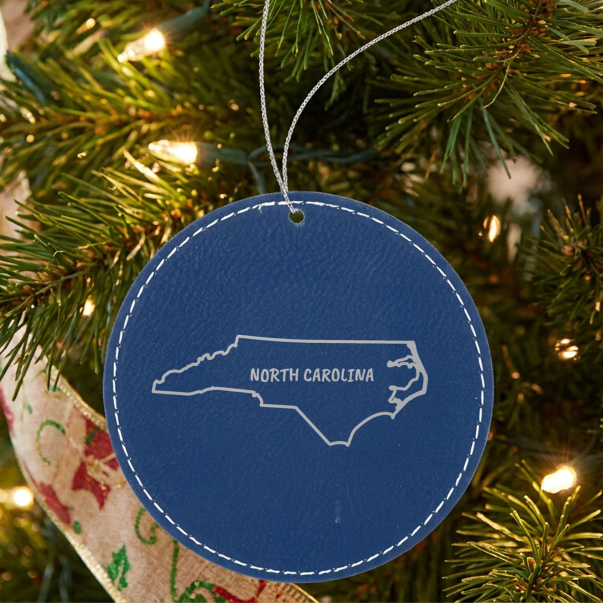 Blue leatherette round Christmas ornament laser engraved with the North Carolina Brush Lettering design, hanging on a tree